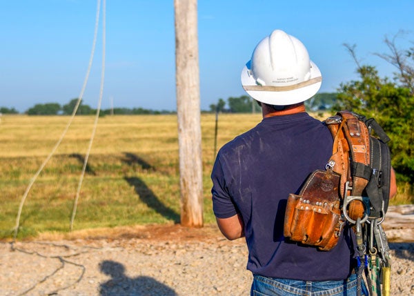 A lineman stands with his back to the camera looking at a power pole. He is wearing his safety hard hat which says "Safely Home, Everyone, Everyday" and has his climbing belt slug over his right shoulder.